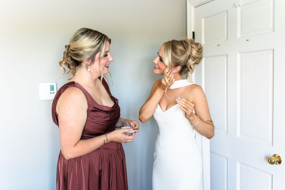 Bride in white gown adjusting earring while bridesmaid in mauve dress holds jewelry box during wedding preparations.