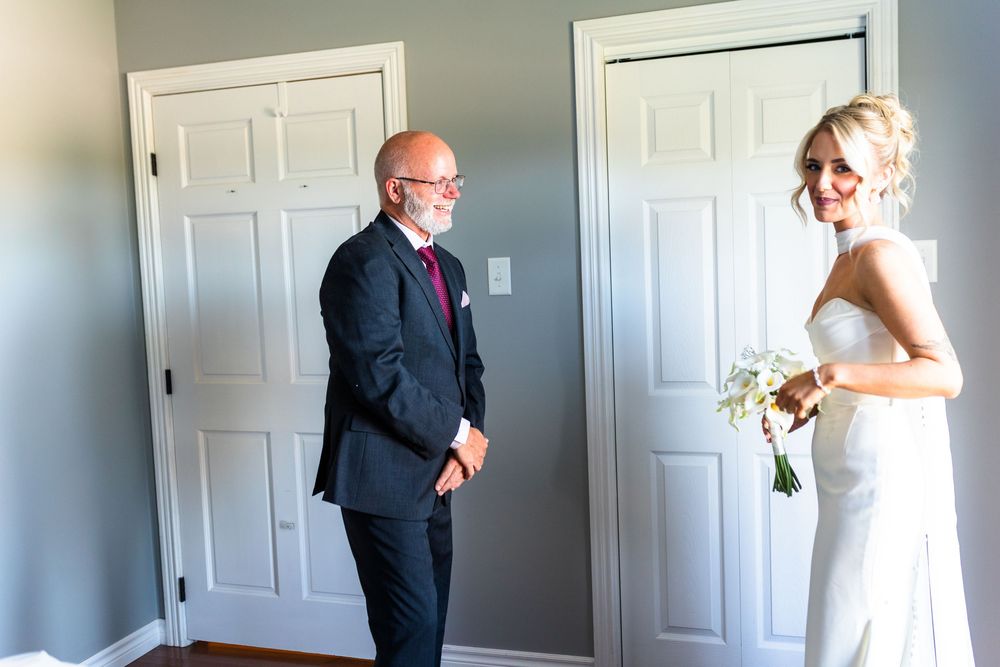 A bride in a white gown holds a bouquet while an older man in a dark suit smiles at her indoors.