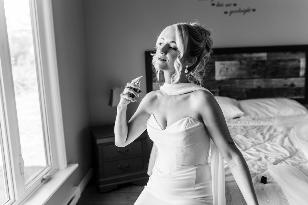 Bride in white wedding gown applying perfume by window in black and white photo.