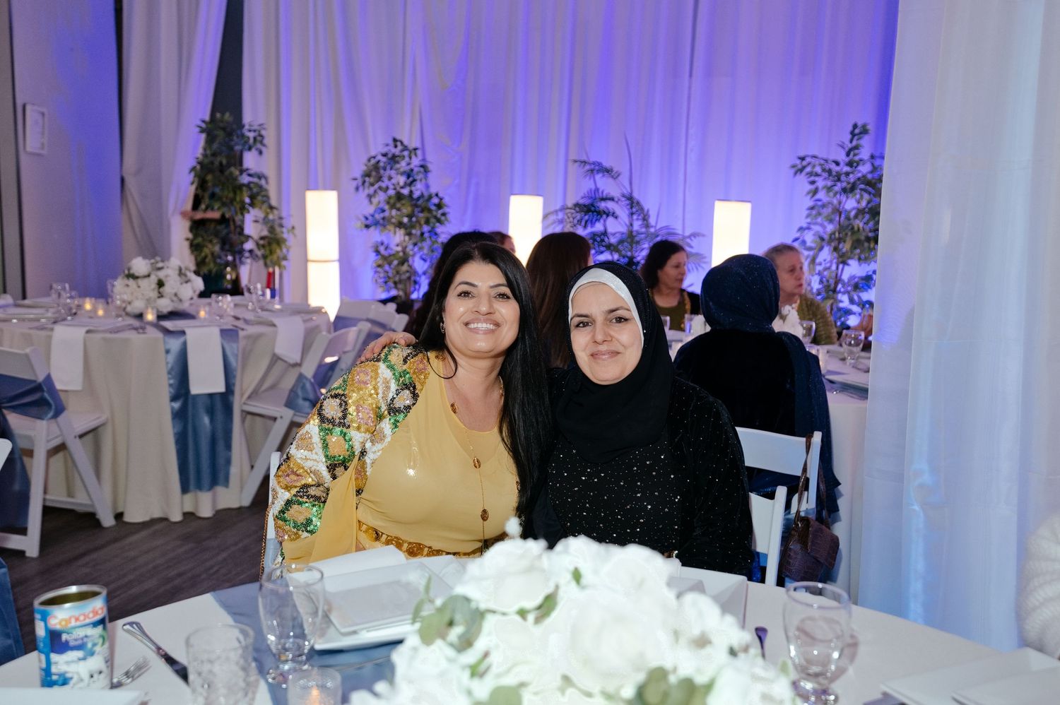 Two smiling women pose close together at a banquet table decorated with white floral centerpieces, with guests and draped decor behind them.