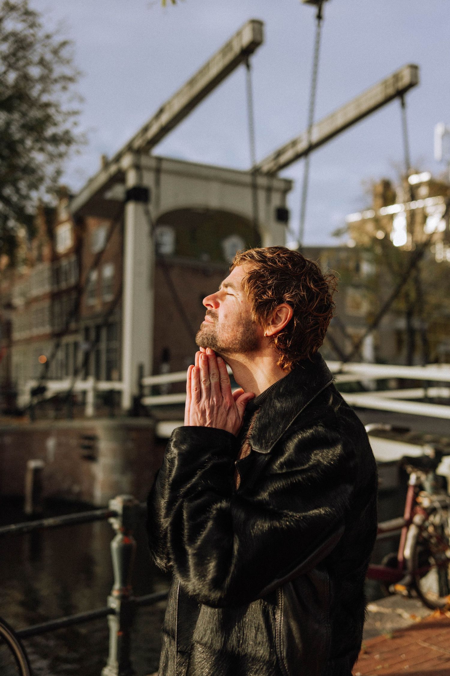 Man in a black fur coat with hands pressed together and eyes closed beside a canal, with a historic drawbridge behind him. Amsterdam