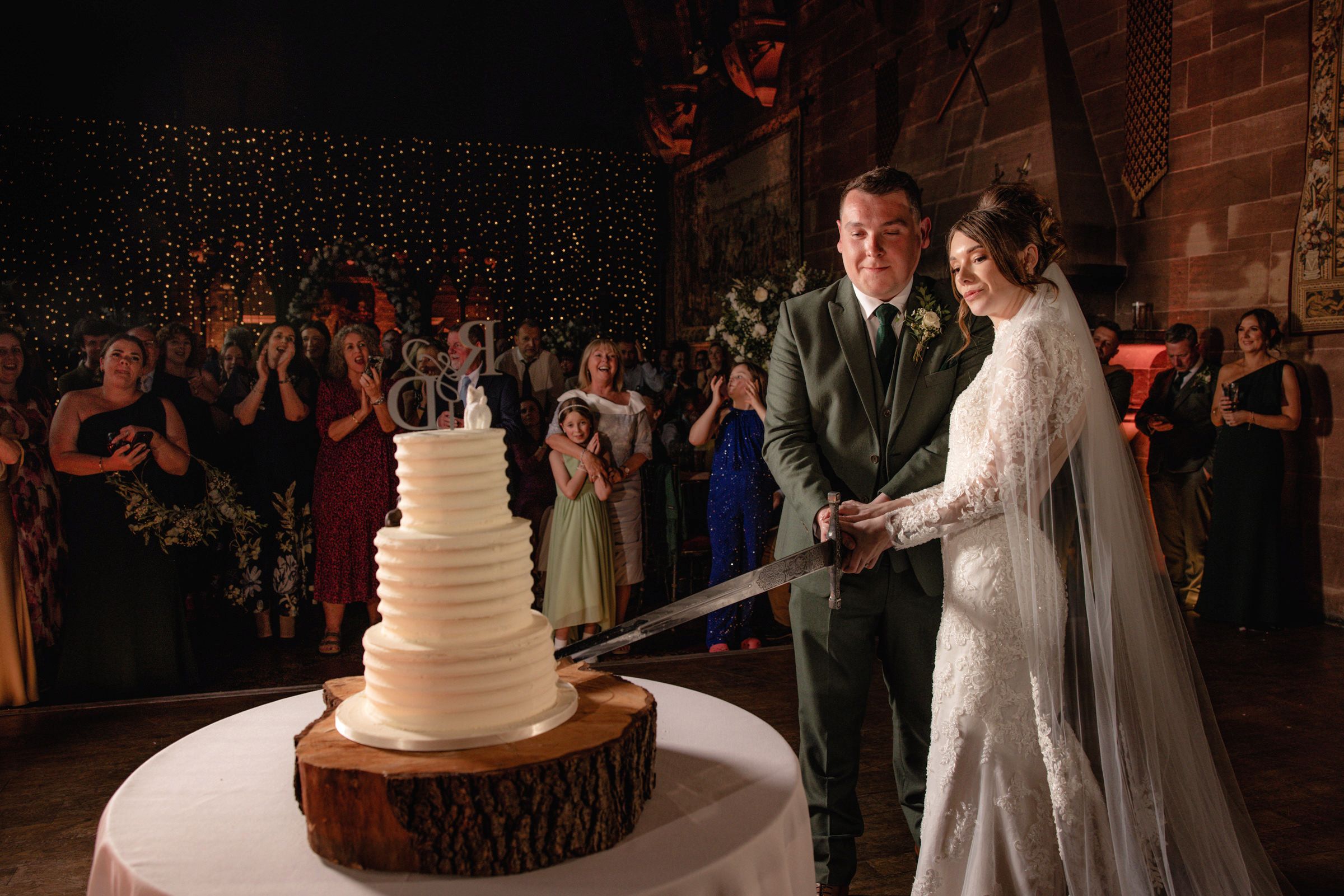 Bride in a lace gown and veil and groom in a green suit cut a white tiered wedding cake on a wooden stand as guests watch.