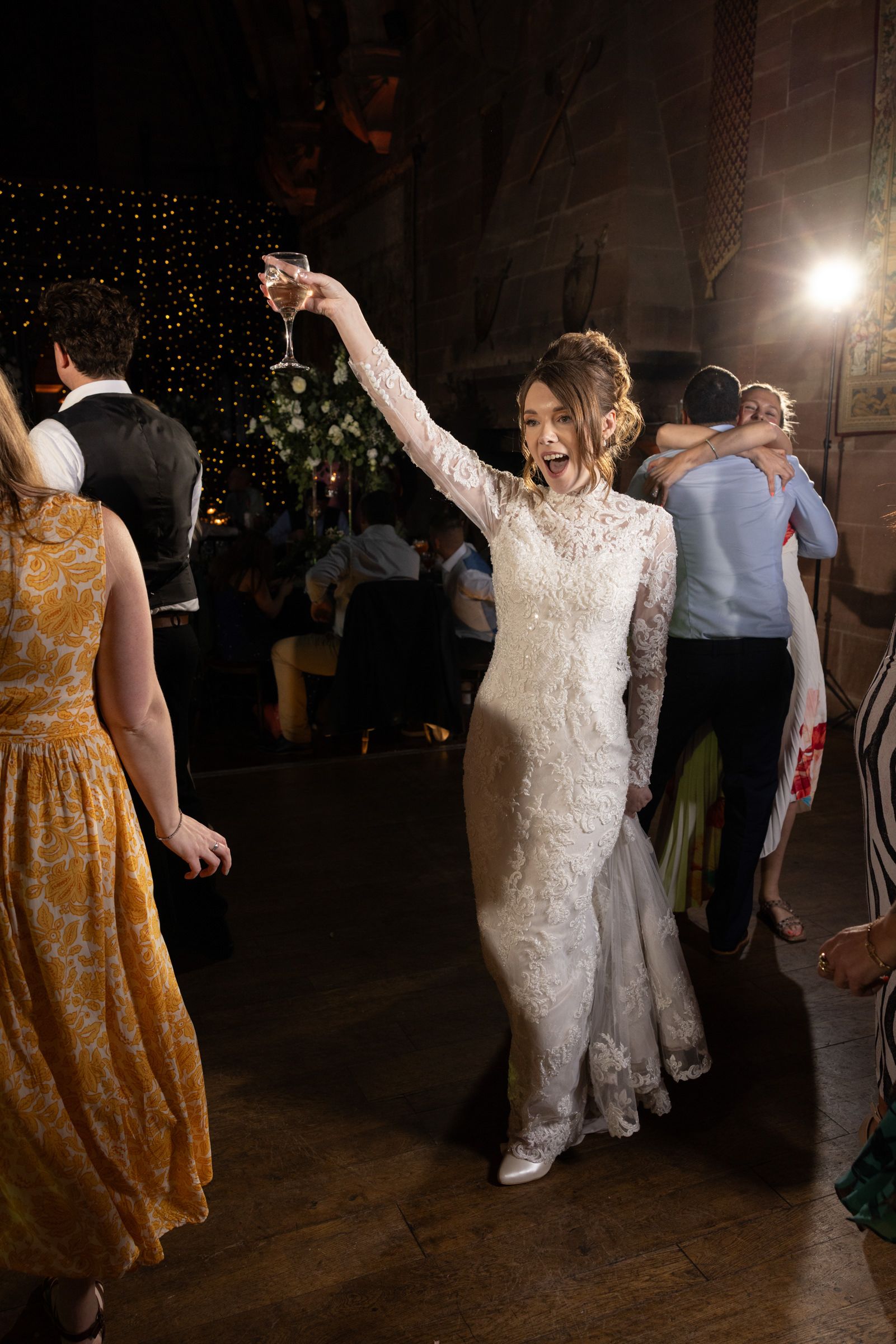 Joyful bride in a white lace gown raising a champagne glass while dancing with wedding guests at an evening reception.