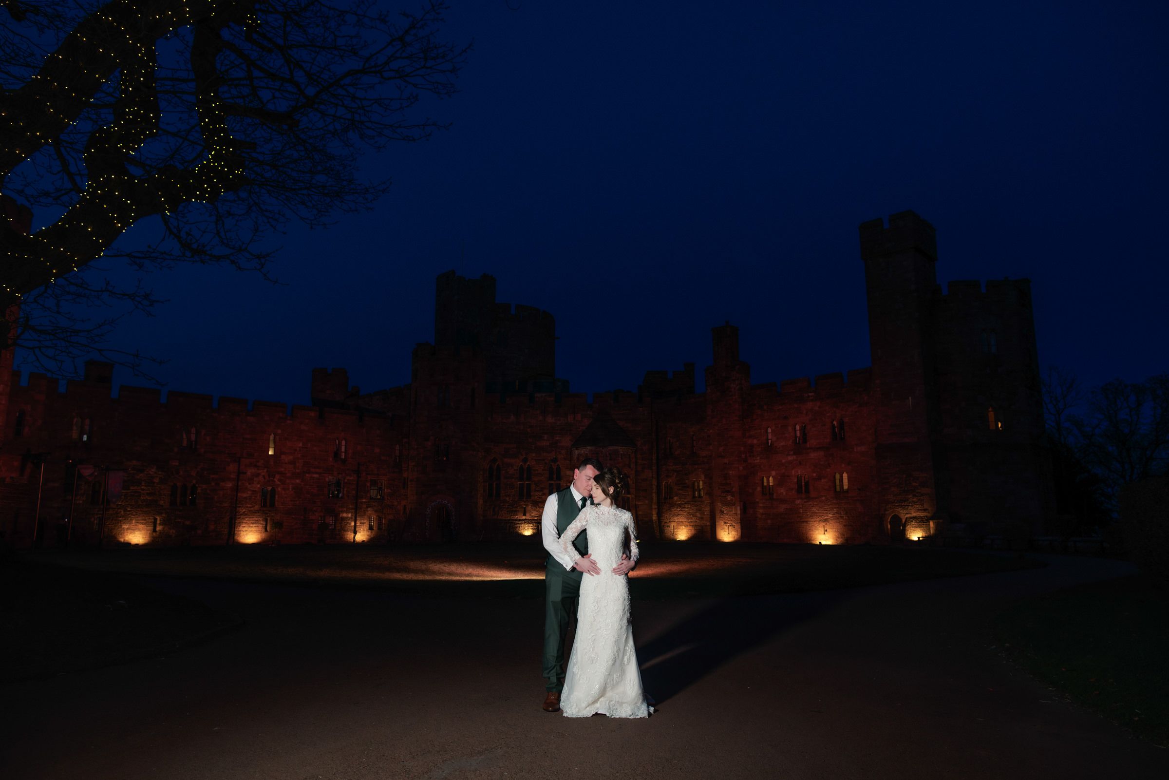 Bride in a white gown and groom in a suit embrace on a lawn at night in front of an illuminated historic castle under a deep blue sky.