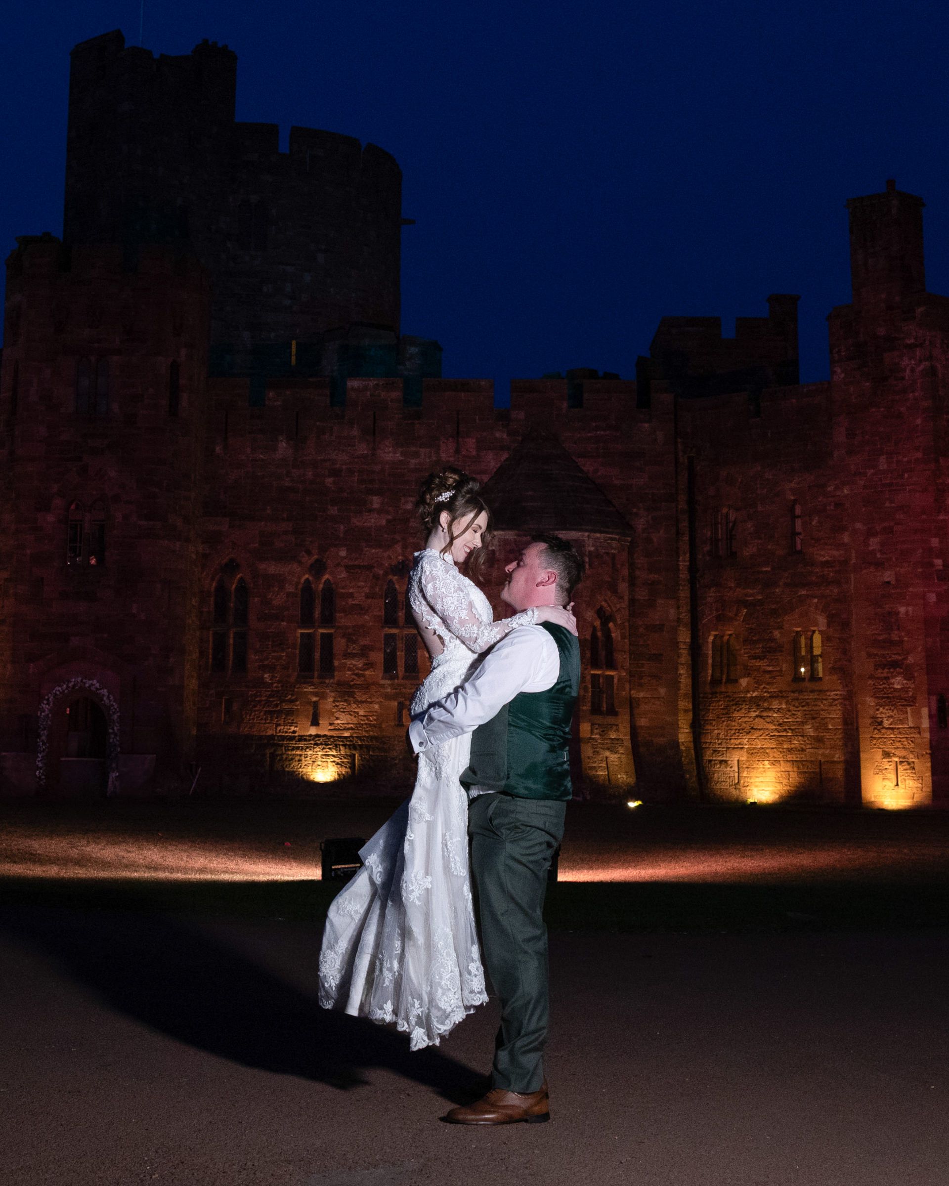 Groom lifting his bride in a white gown for a romantic night embrace in front of an illuminated red stone castle under a deep blue sky.