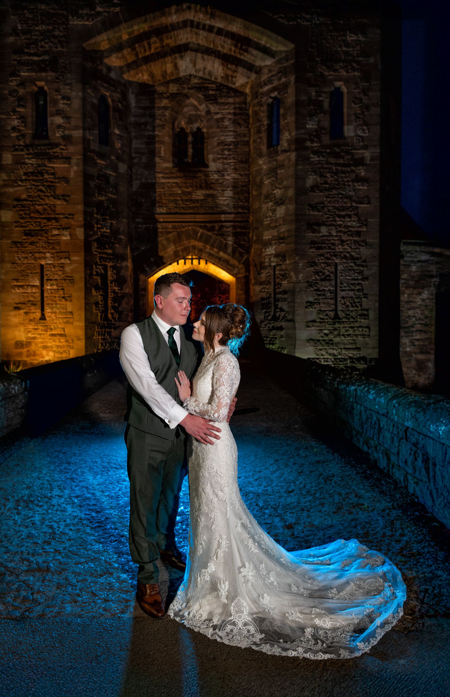 Bride in a lace wedding gown and groom in a green vest embrace on a blue-lit path before a glowing stone castle archway at night.