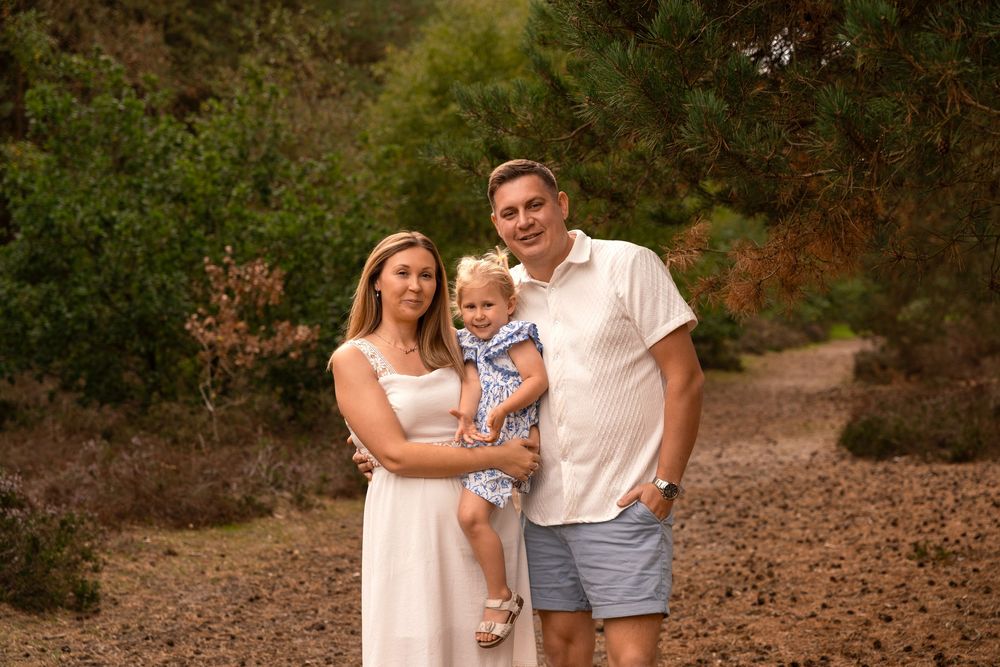 Smiling family of three posing outdoors in a sunny wooded area, with a mother in a white dress, a father in a polo, and a toddler girl.