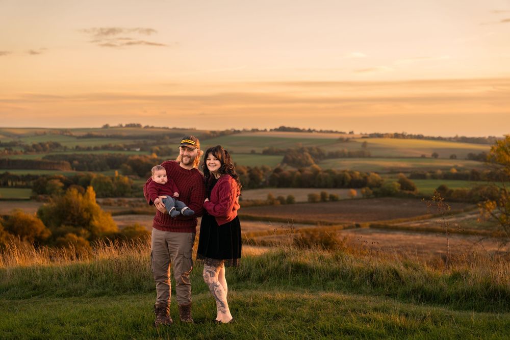 Family of three, a man, woman and toddler, posing on a grassy hilltop overlooking rolling countryside at golden hour sunset.
