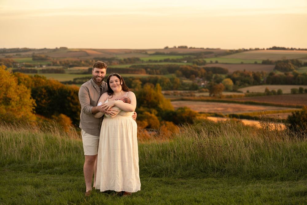 Smiling couple embracing in a grassy field at sunset, the woman in a flowing cream dress, with rolling countryside hills behind them.