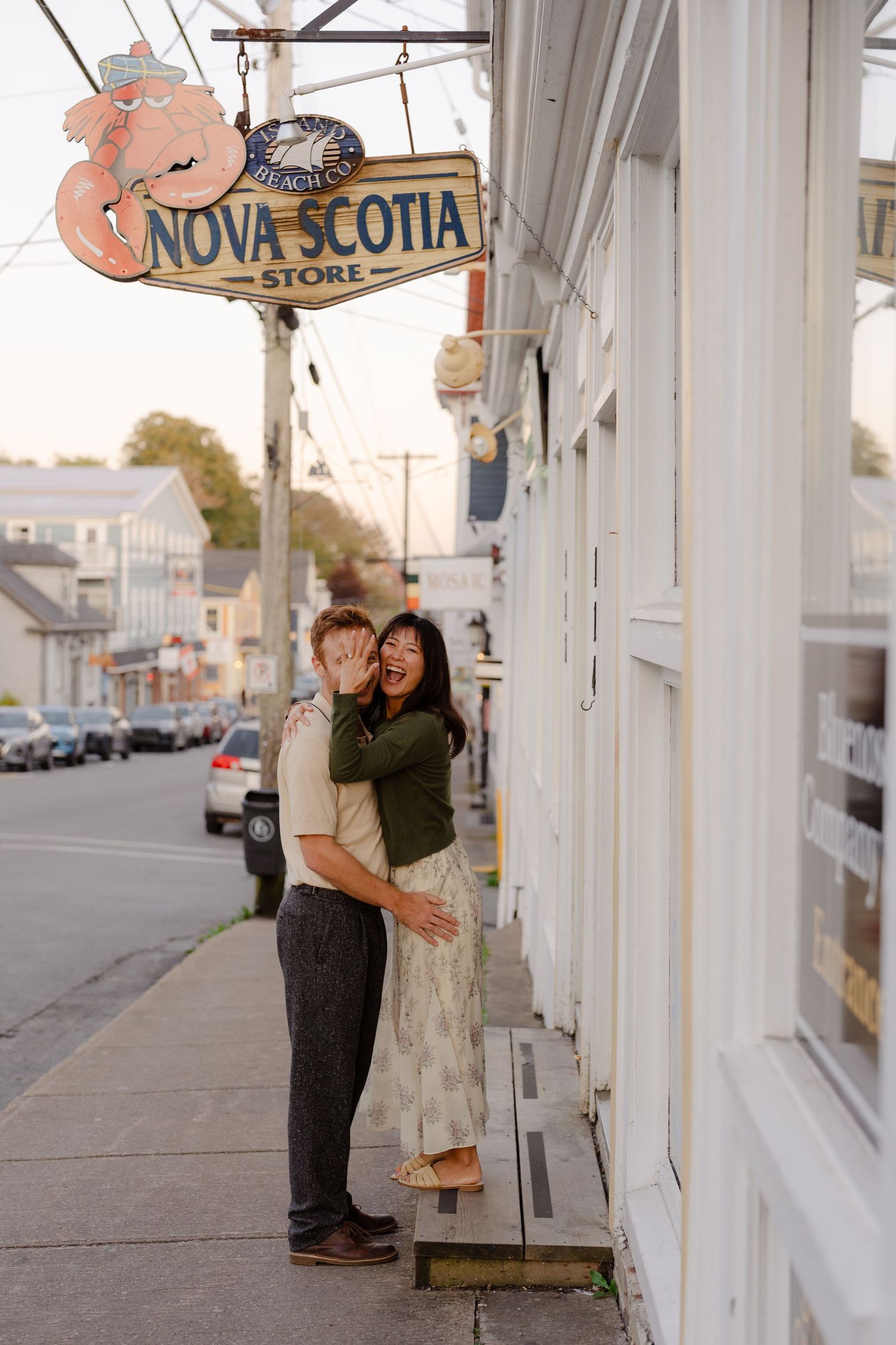 Couple embracing and laughing under a Nova Scotia Store sign on a charming Lunnenberg sidewalk at golden hour.