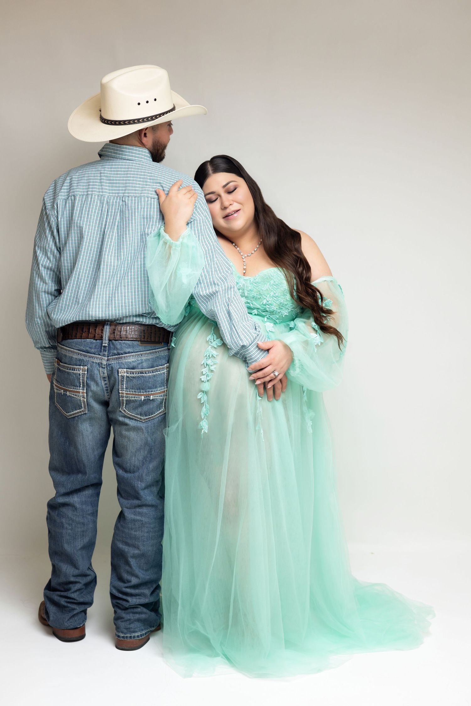 Pregnant woman in mint green tulle gown poses with man in cowboy hat and jeans in a maternity studio portrait.