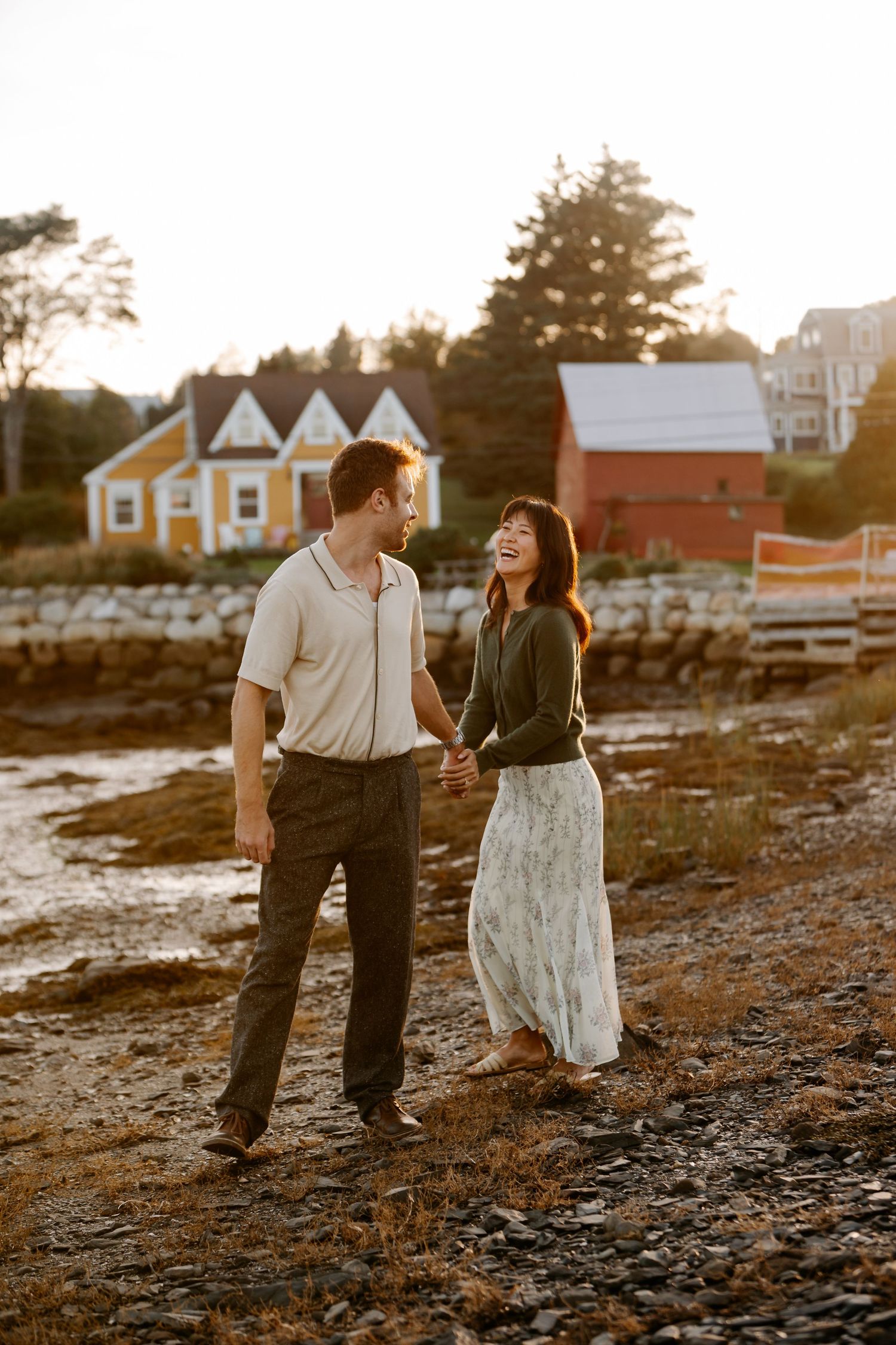 Couple holding hands walking along rocky shoreline at golden hour with colorful Lunnenberg coastal buildings behind them.
