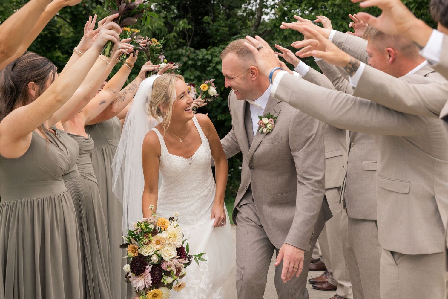 Bride and groom walk through arch of raised hands formed by wedding party in sage bridesmaid dresses and tan suits in Ohio