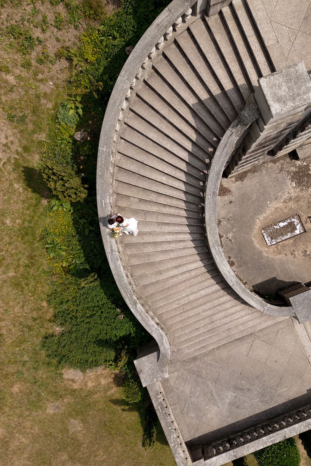 Aerial view of a curved concrete staircase with two people standing on the steps, surrounded by green grass and shrubs.