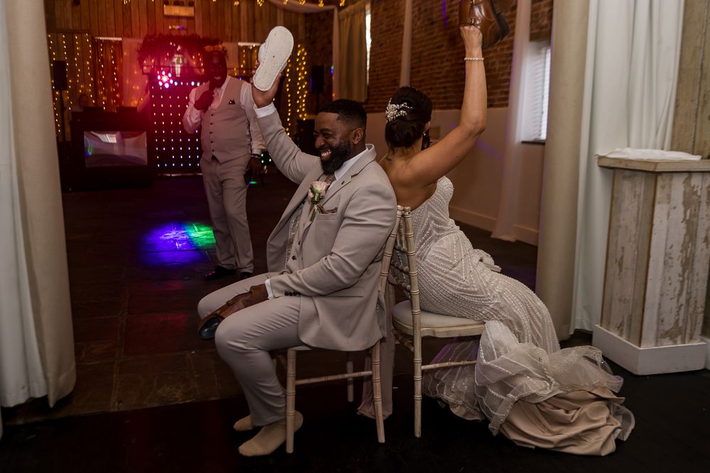 Bride and groom sitting back-to-back on chairs, raising hands joyfully during wedding reception dance floor celebration.