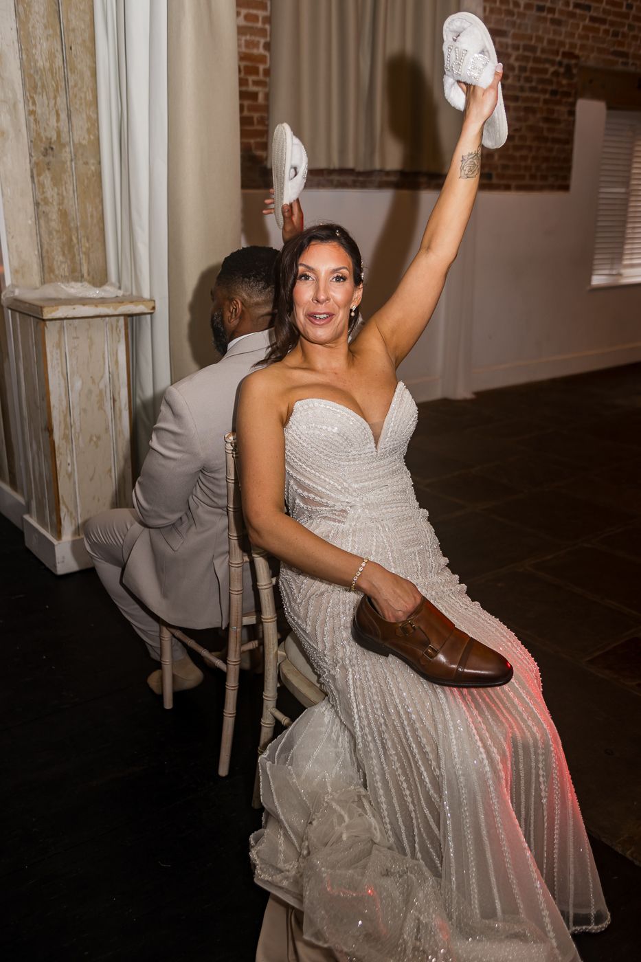 Bride in sparkly gown holds white shoes aloft at wedding reception, sitting beside groom in gray suit.