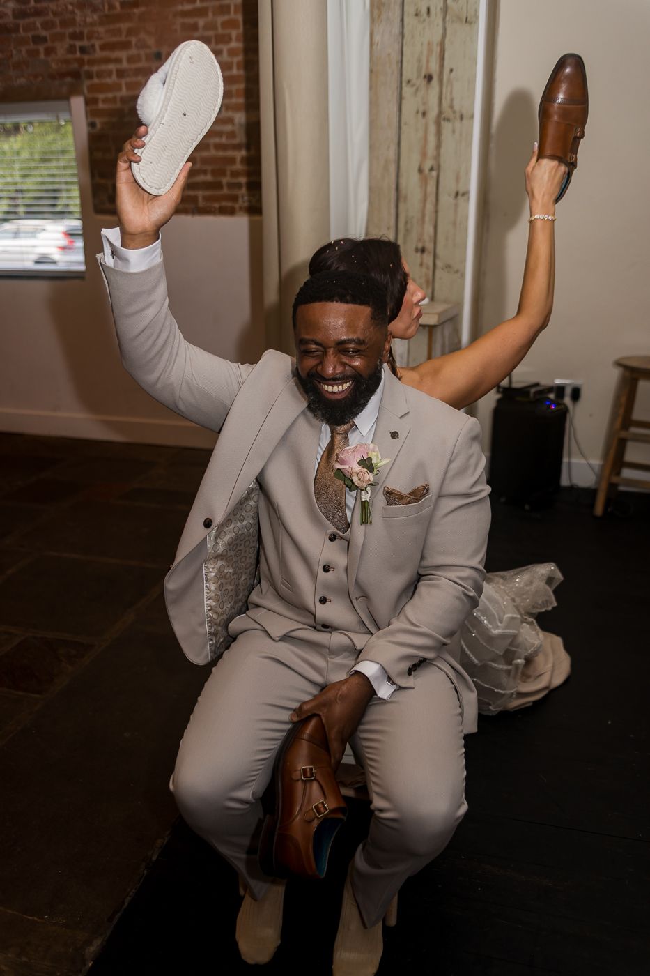 Joyful groom in beige suit holds shoes aloft during fun wedding shoe game ceremony.