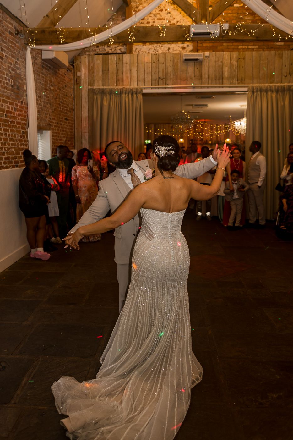 Bride and groom share their first dance in a rustic barn wedding venue with fairy lights and guests watching.