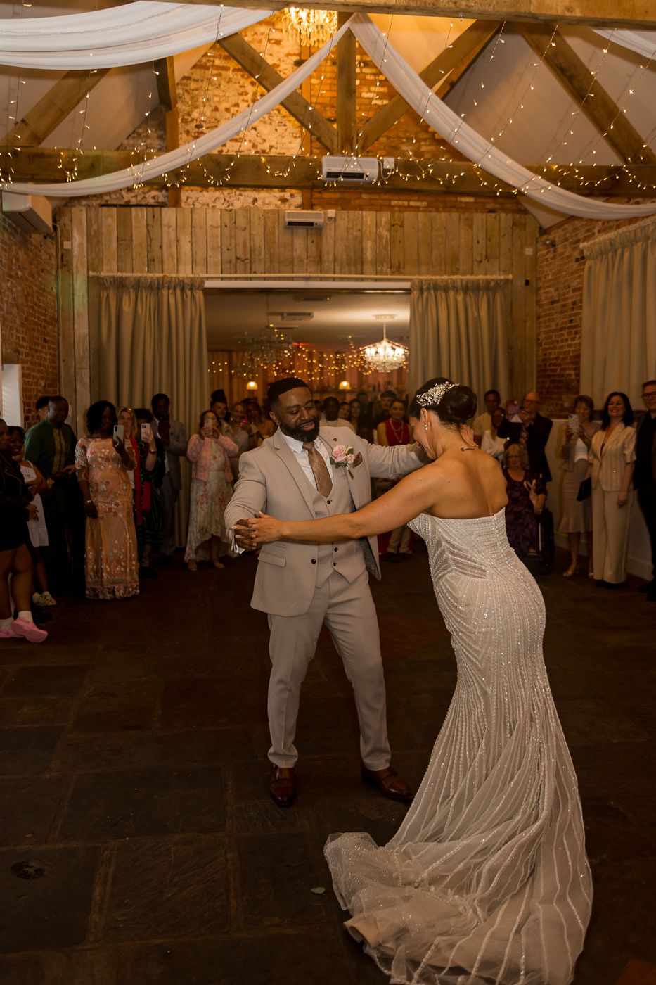 Bride and groom share their first dance in a rustic barn wedding venue decorated with fairy lights and draped fabric.