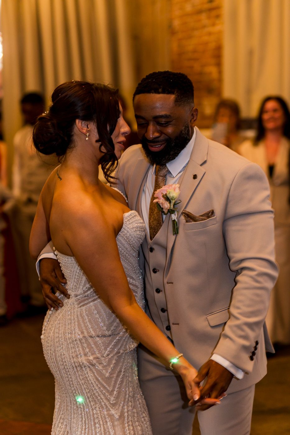 Bride and groom share first dance, groom in tan suit with boutonniere, bride in beaded gown, wedding reception.