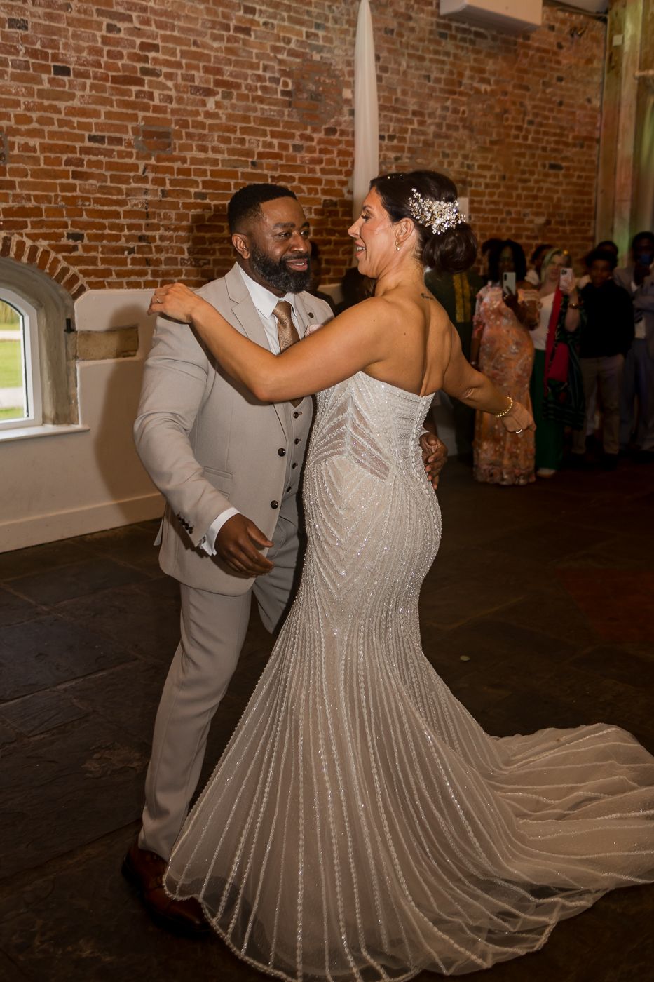 Bride and groom share their first dance in a rustic brick venue, bride in beaded gown, groom in tan suit.