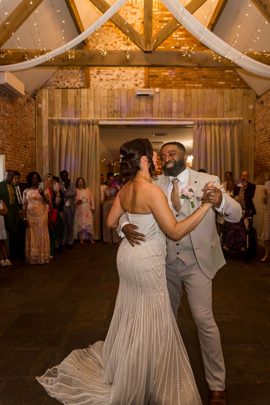Bride and groom share their first dance in a rustic barn wedding venue with exposed brick walls and fairy lights.
