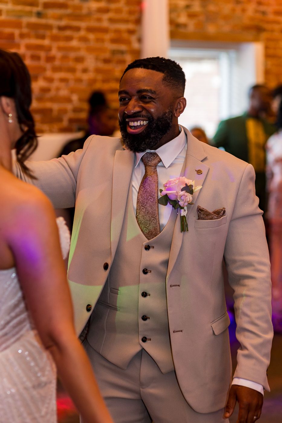 A smiling groom in a beige three-piece suit with floral boutonniere dances at a wedding reception.