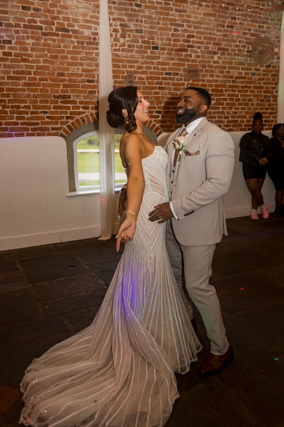Bride and groom share first dance in elegant wedding attire against rustic brick wall venue.