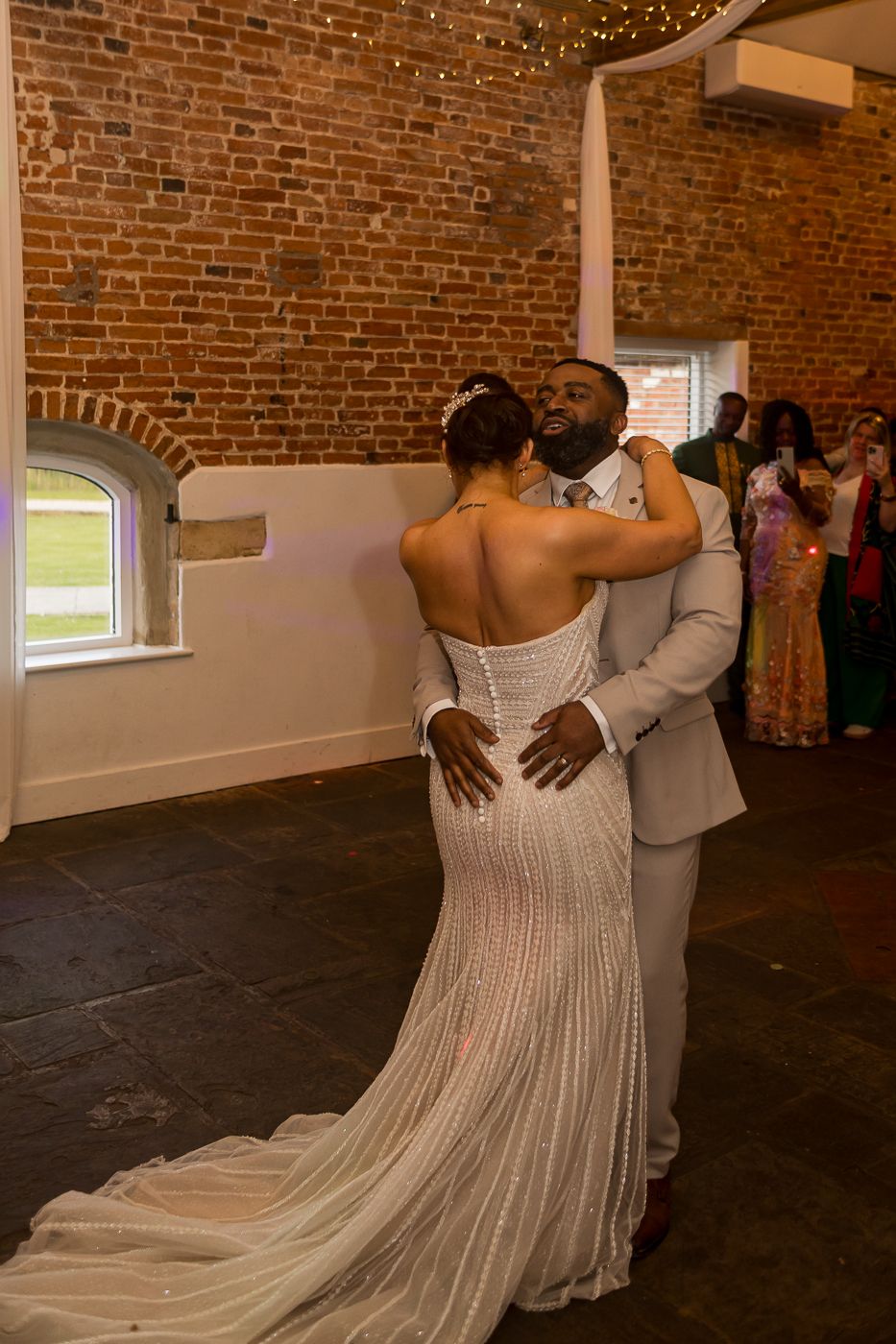 Bride and groom share their first dance in a rustic brick-walled venue, guests watching in the background.