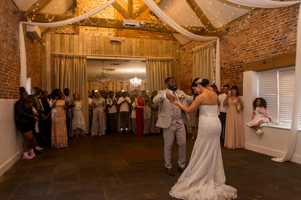 Bride and groom share first dance in rustic brick barn wedding venue surrounded by guests.