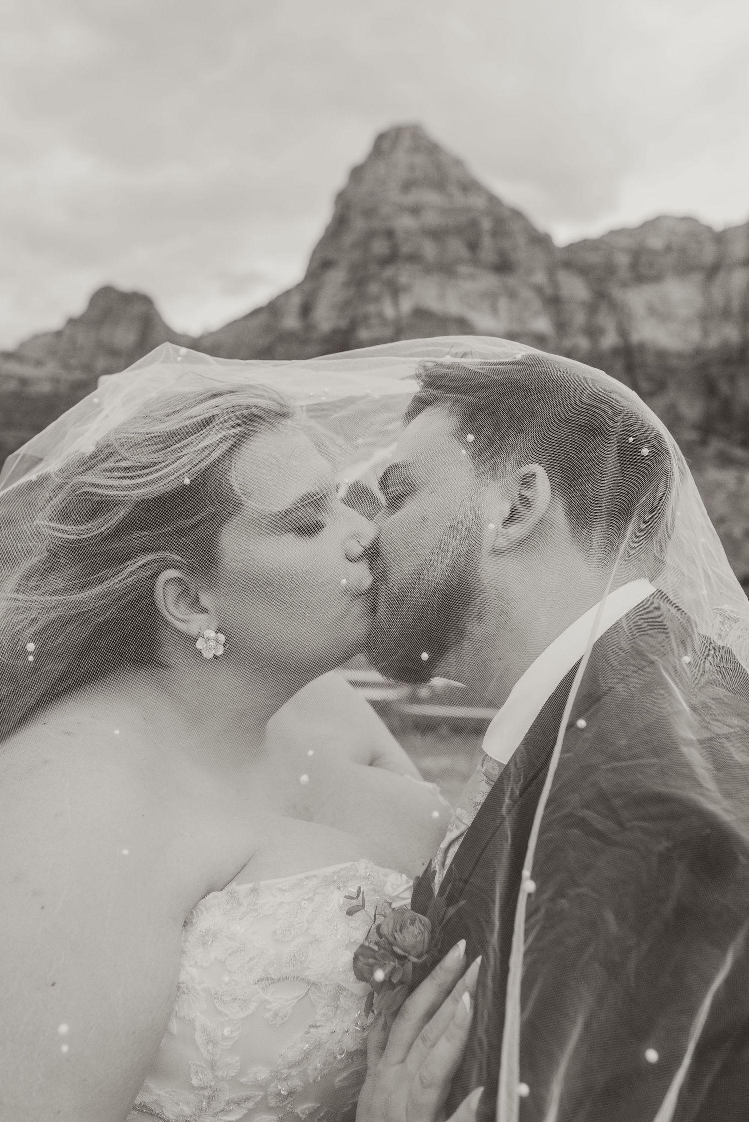 Black and white photo of bride and groom kissing under veil with mountain backdrop.