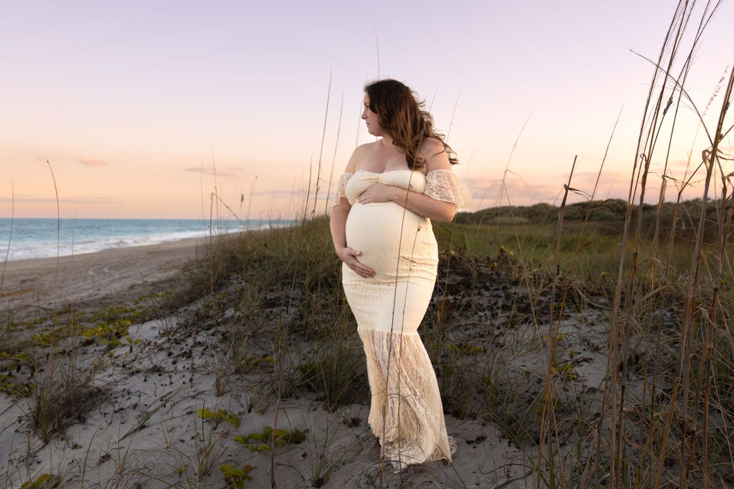 Pregnant woman in a flowing cream lace gown cradles her baby bump on a sandy dune with sea oats at sunset beside the ocean.
