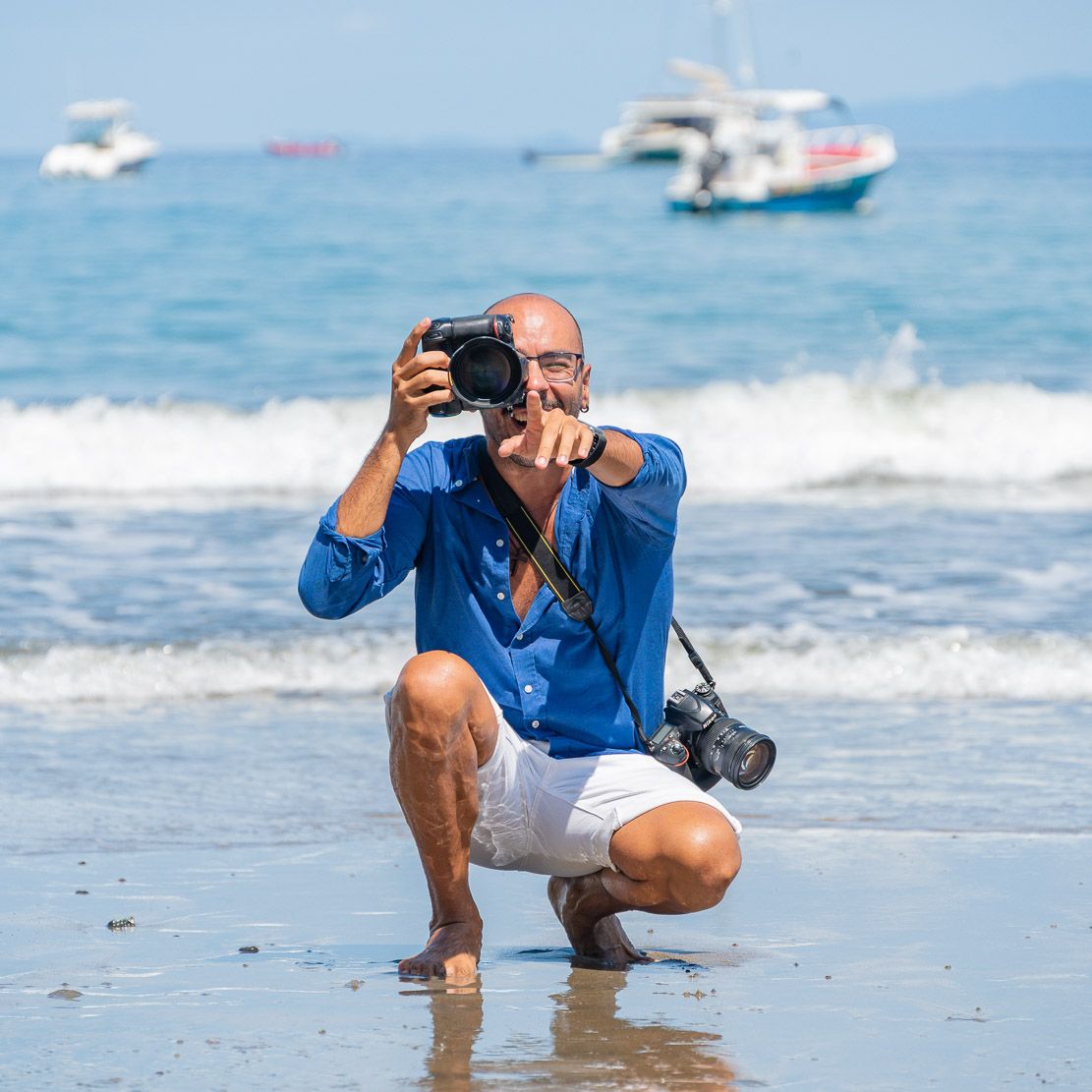 Bald man in a blue shirt and white shorts crouches on a wet beach, taking a photo with a camera as boats float behind the waves.