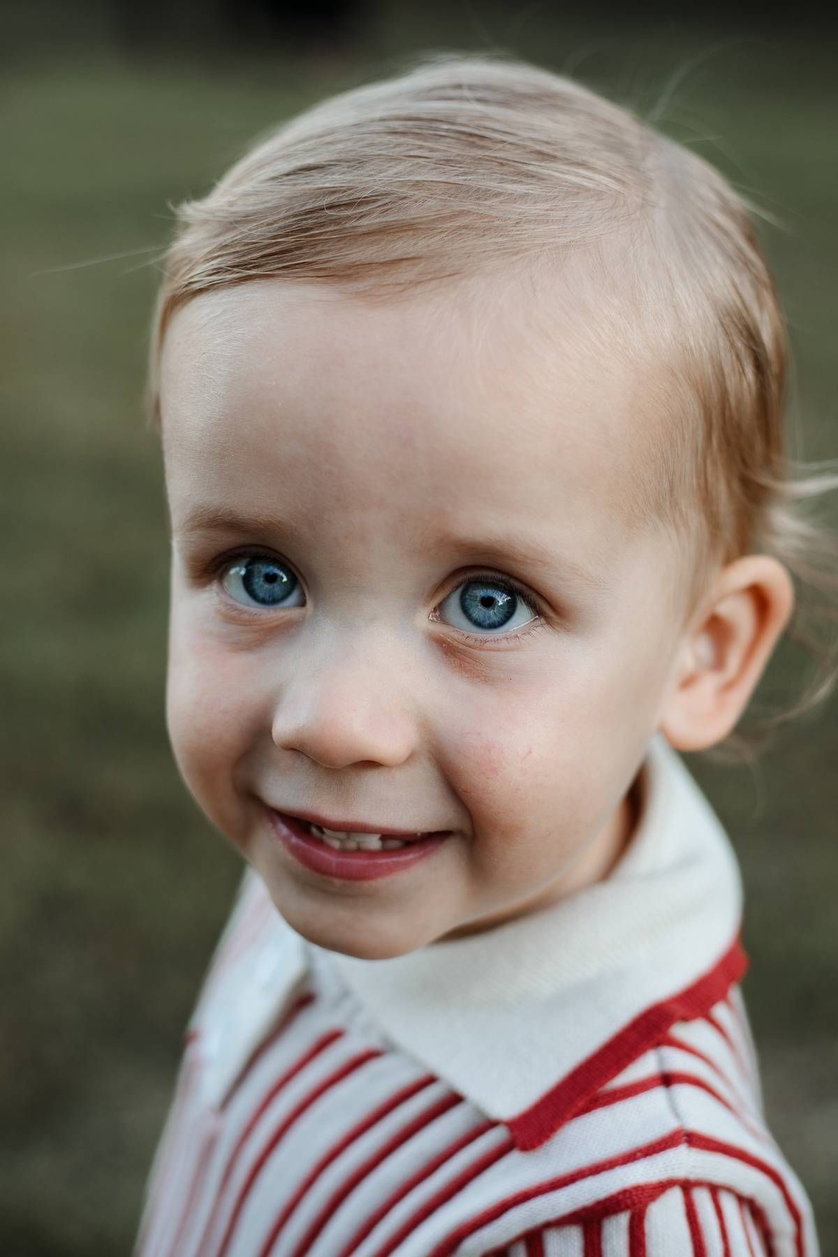 Close-up portrait of a smiling blue-eyed toddler with light hair wearing a red and white striped collared shirt outdoors.
