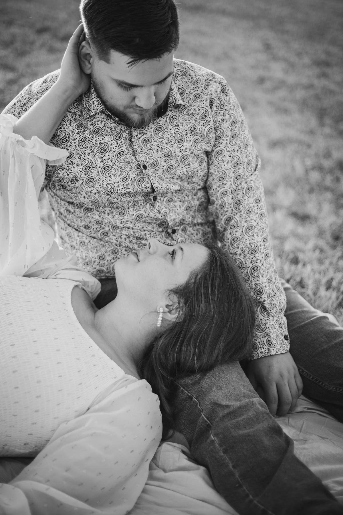Black and white photo of a bearded man in a patterned shirt gazing down at a woman resting her head in his lap outdoors.