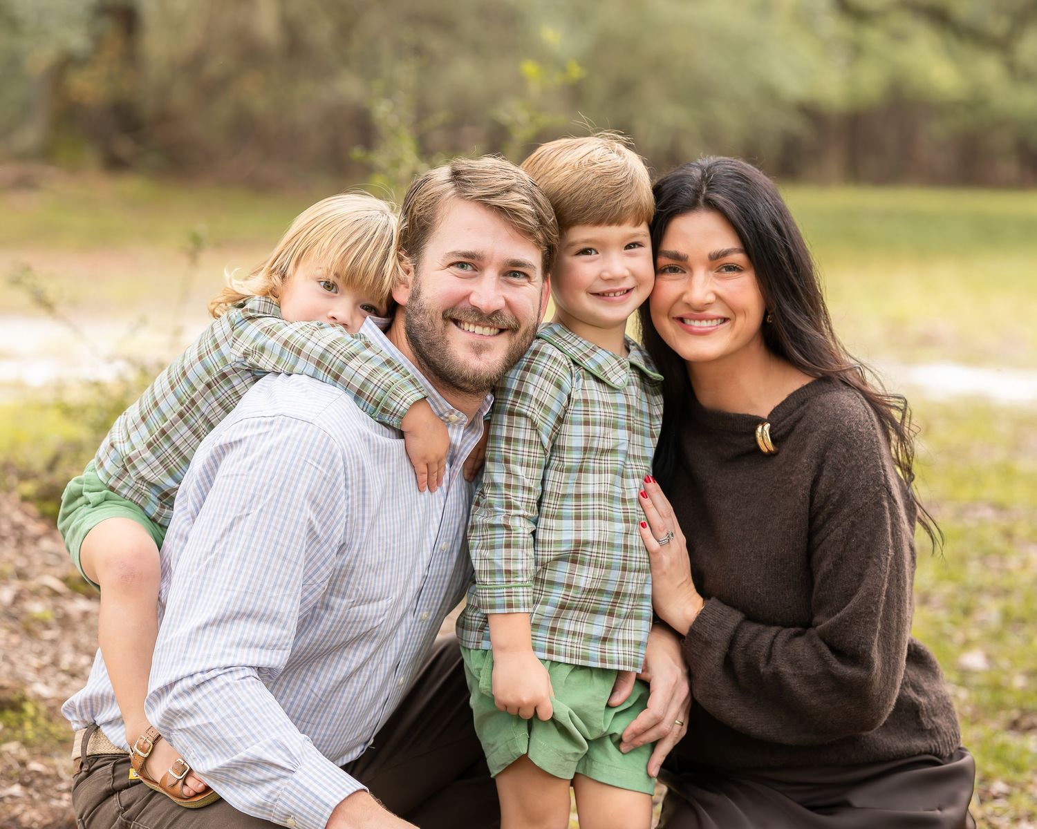A smiling family of four poses outdoors, with a father and mother embracing two young boys in matching plaid shirts on a grassy field.
