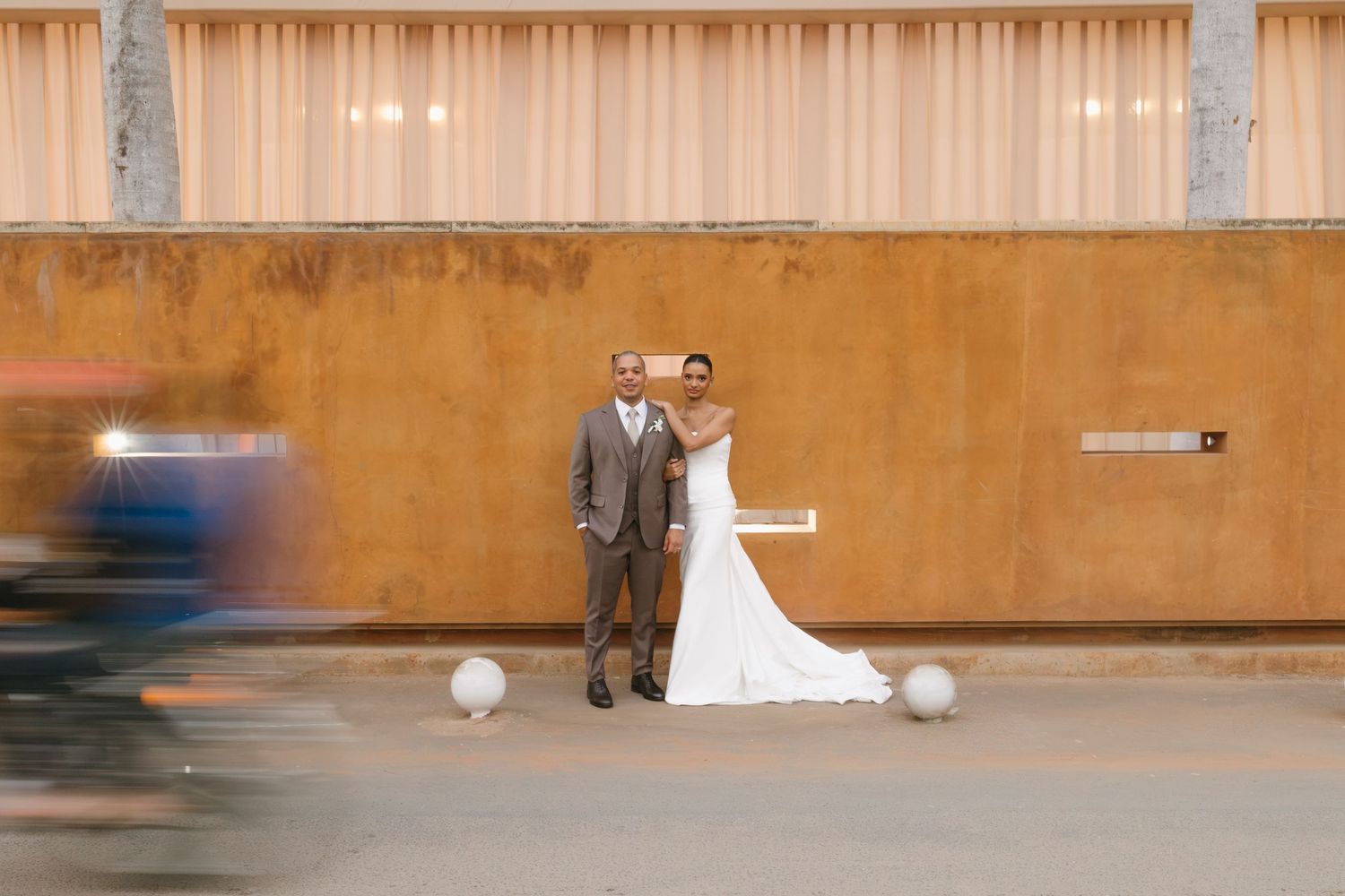 Bride in white gown and groom in brown suit pose against rustic orange wall outdoors on their wedding day.