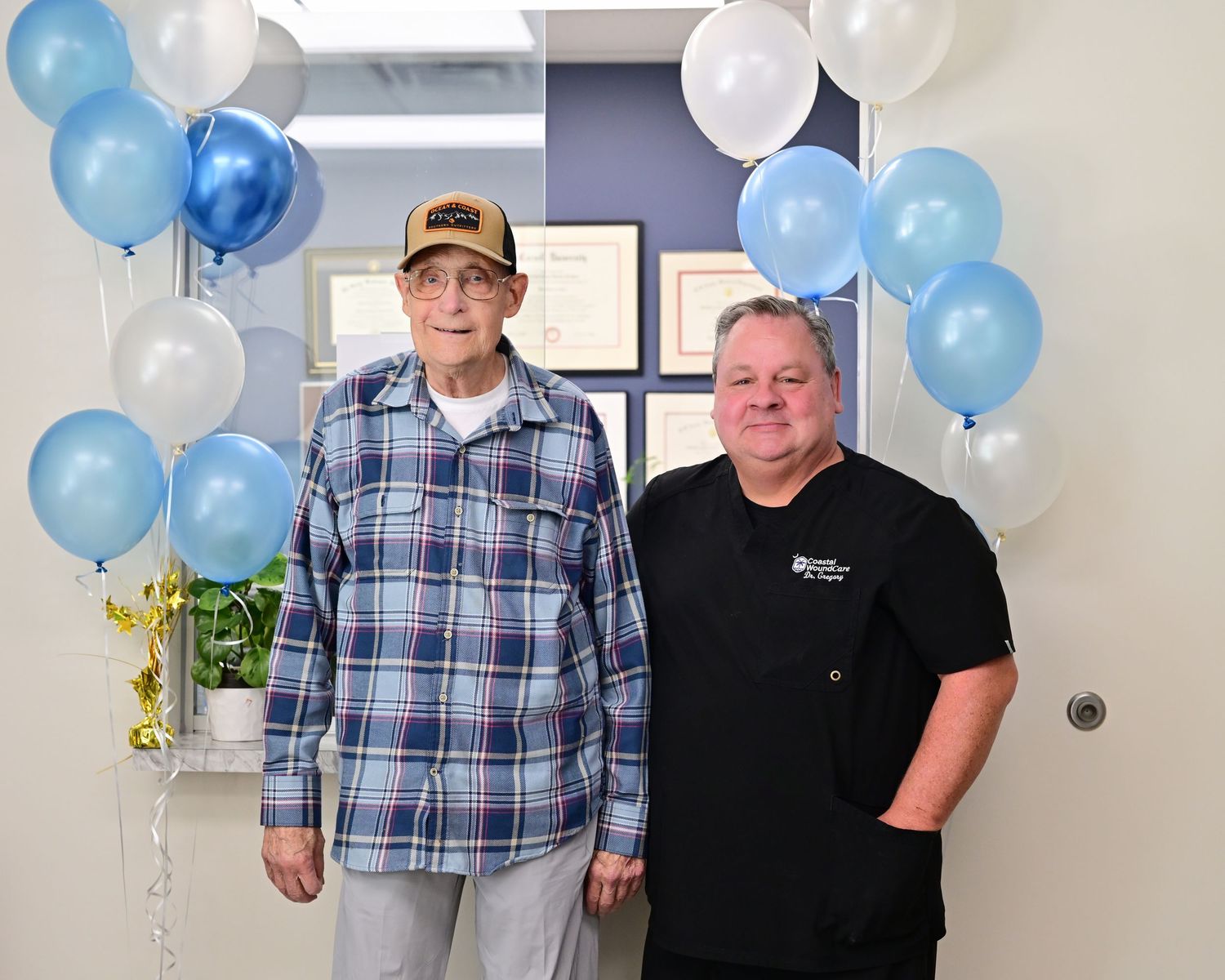 Two men pose under blue and white balloon arch in medical office celebrating a milestone achievement.