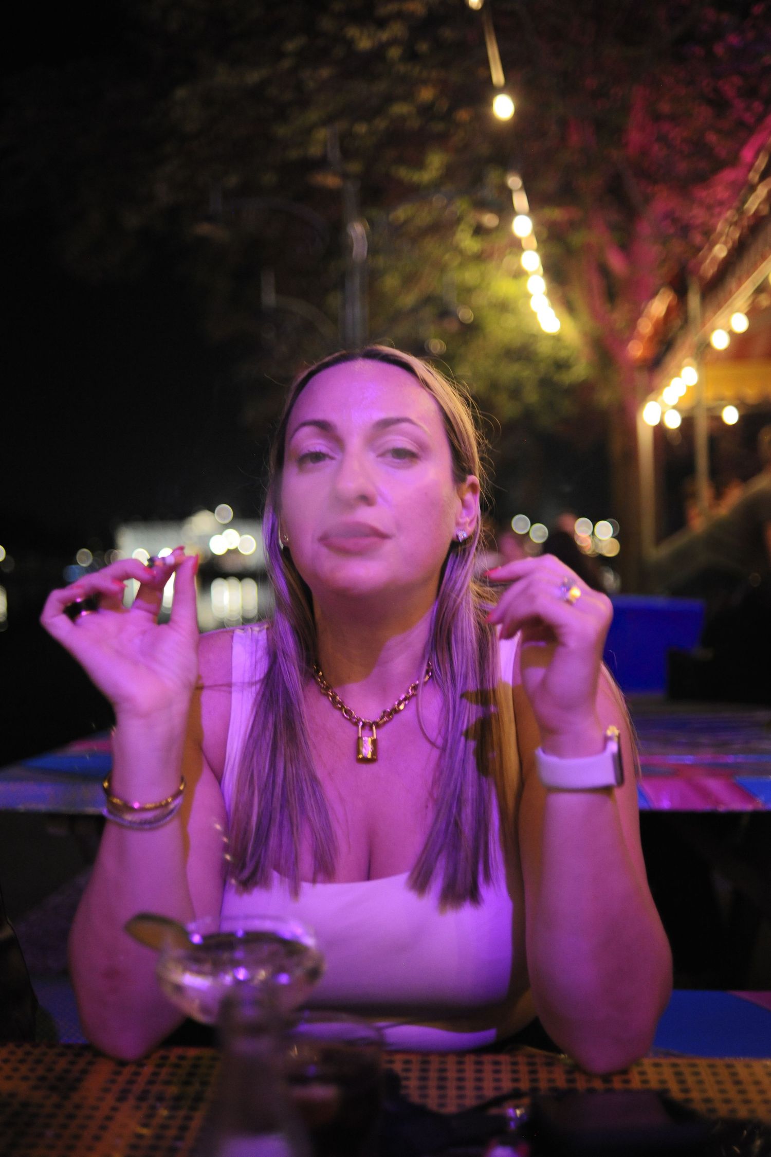 Woman holding a glass of white wine outdoors at night, wearing a floral jacket and gold necklace.