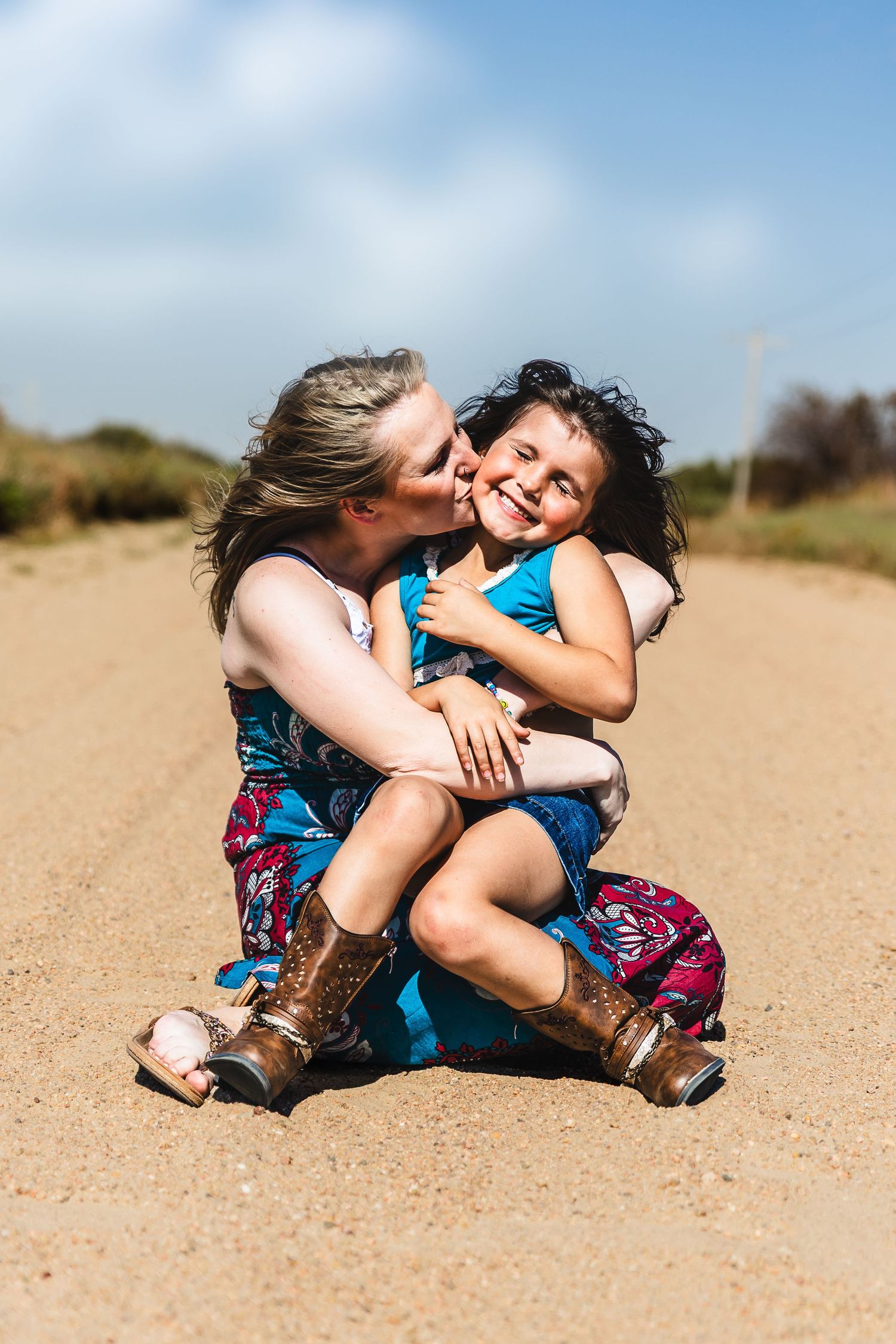 A woman kisses a smiling young girl while sitting together on a sunny dirt road.