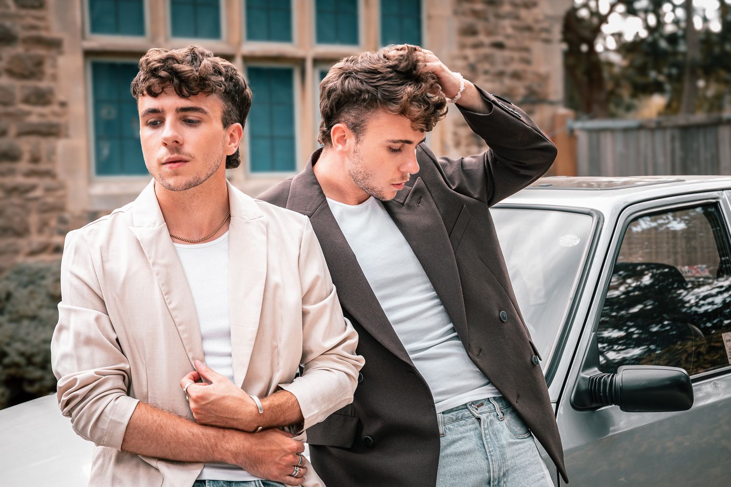 Two young men in blazers pose near a vintage car outside a stone building.