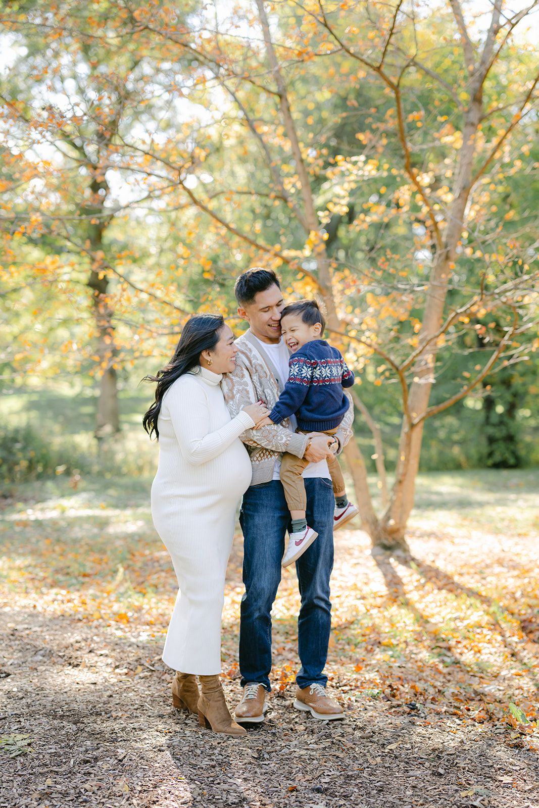 Pregnant woman in white dress with husband holding toddler son during autumn family maternity photo session outdoors.