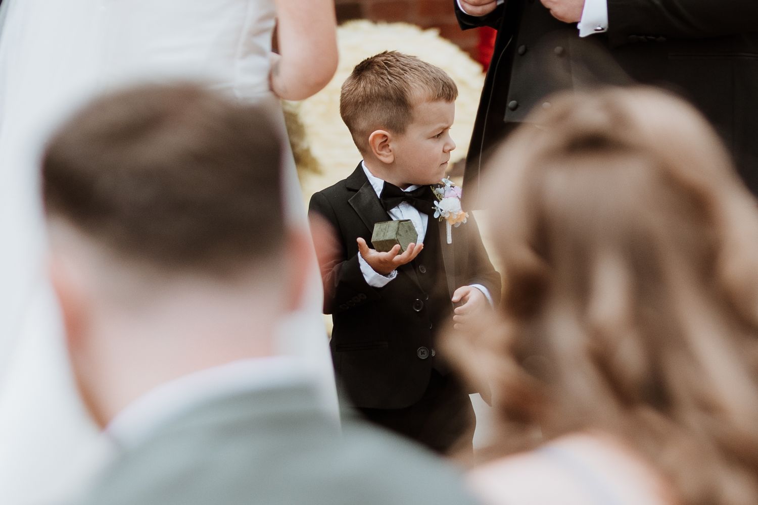 Young ring bearer in black tuxedo and bow tie holds wedding rings during ceremony.