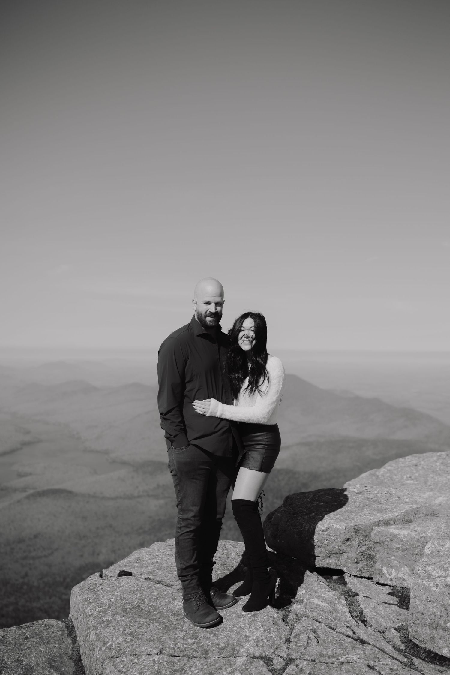 Couple posing on mountain summit in black and white, woman in leather skirt embracing bald man with scenic ridge views.