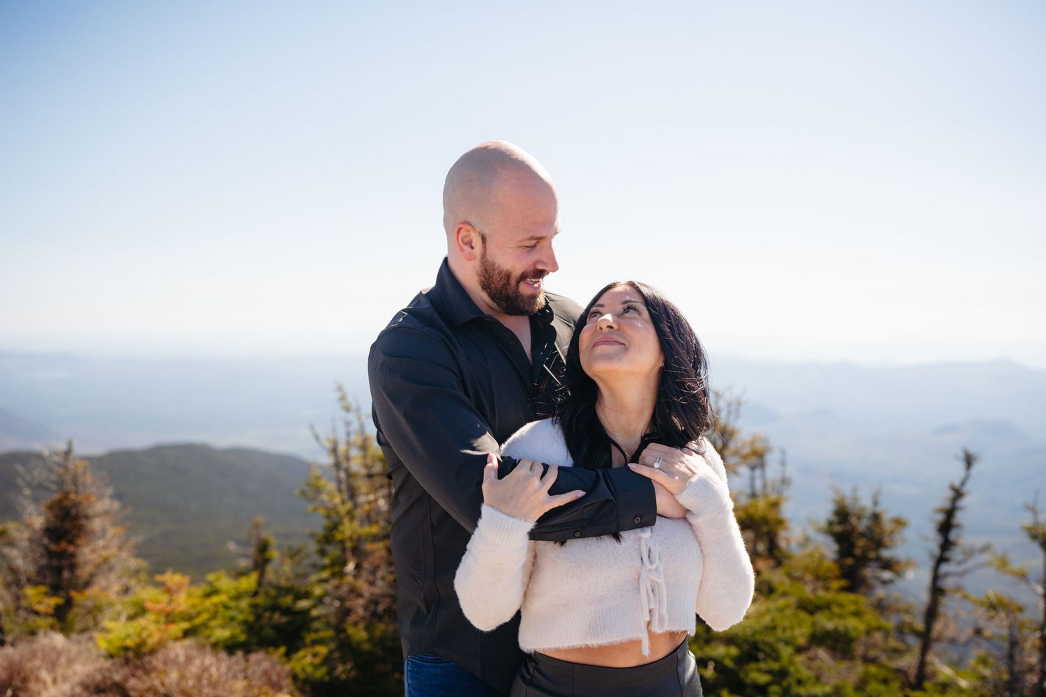 Couple embracing on a mountain summit with scenic blue ridge views on a clear sunny day.