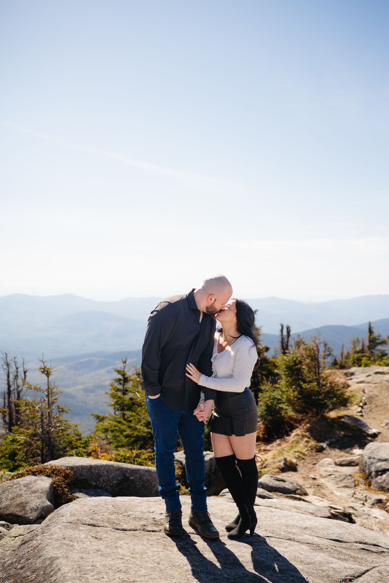 Couple sharing a romantic kiss on a mountain summit with scenic blue ridge views and clear skies.