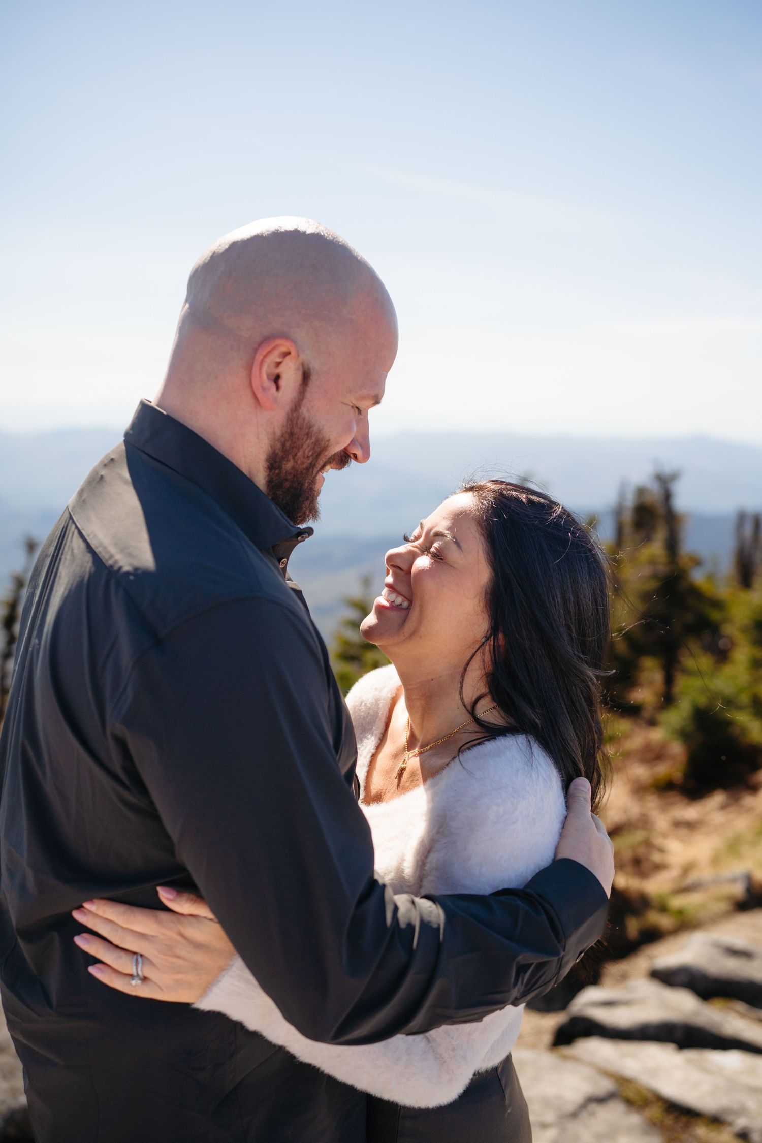 A happy couple embraces and laughs together on a mountain summit, with scenic blue skies and trees in the background.