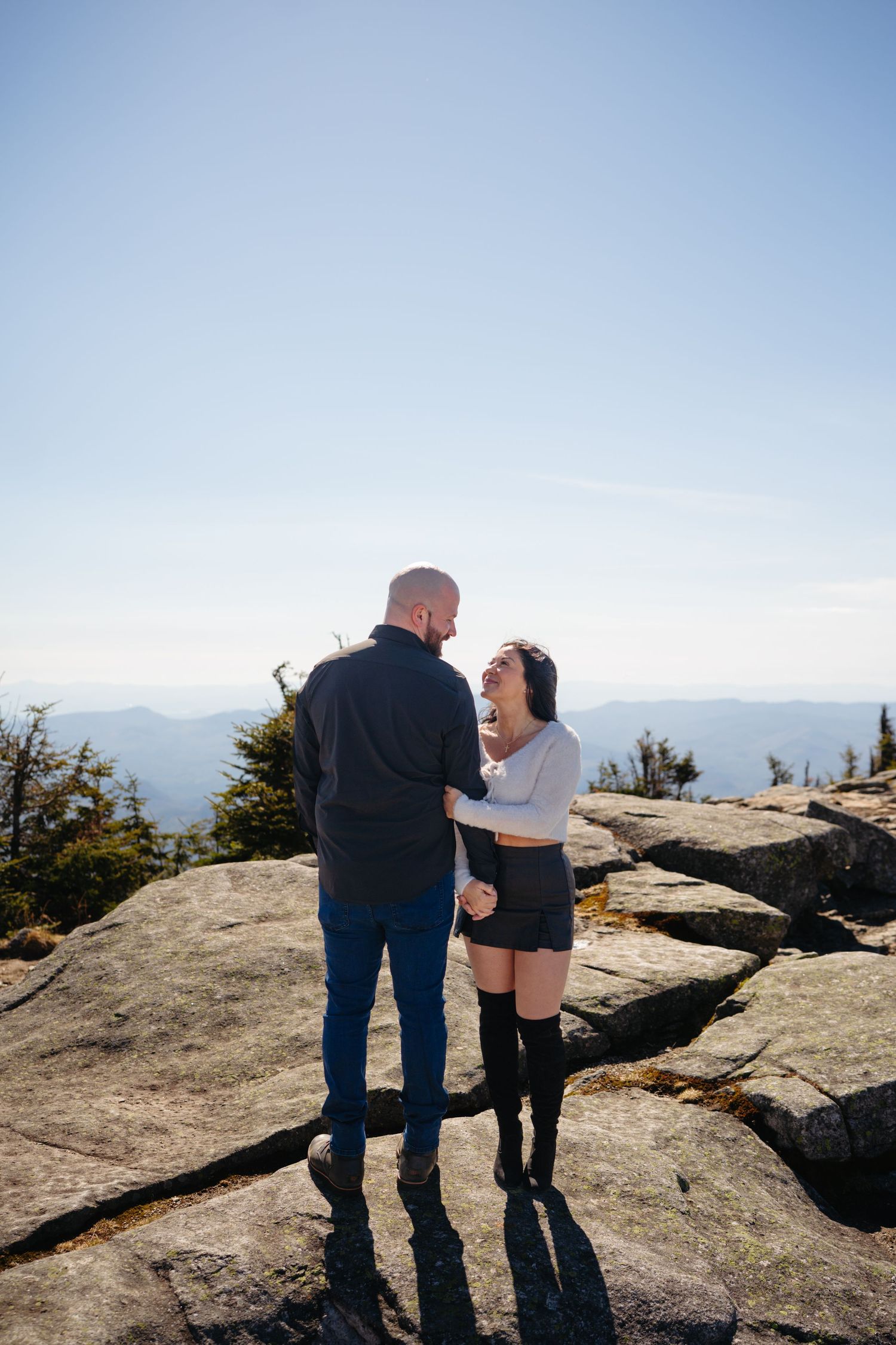 Couple embracing and laughing on rocky mountain summit with scenic blue sky and mountain range backdrop.