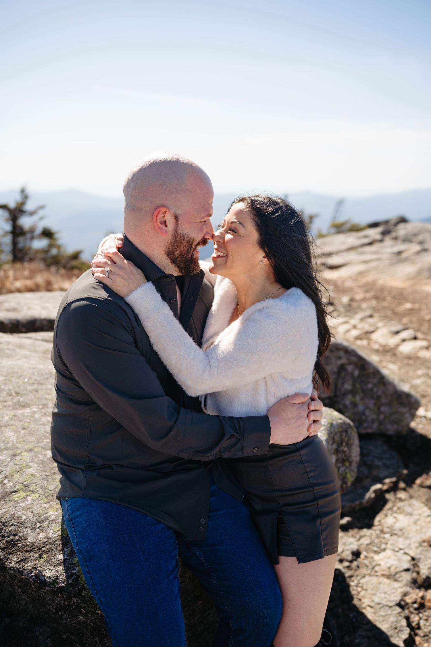 Couple embracing and smiling on a rocky mountain summit with scenic blue sky and mountain range backdrop.