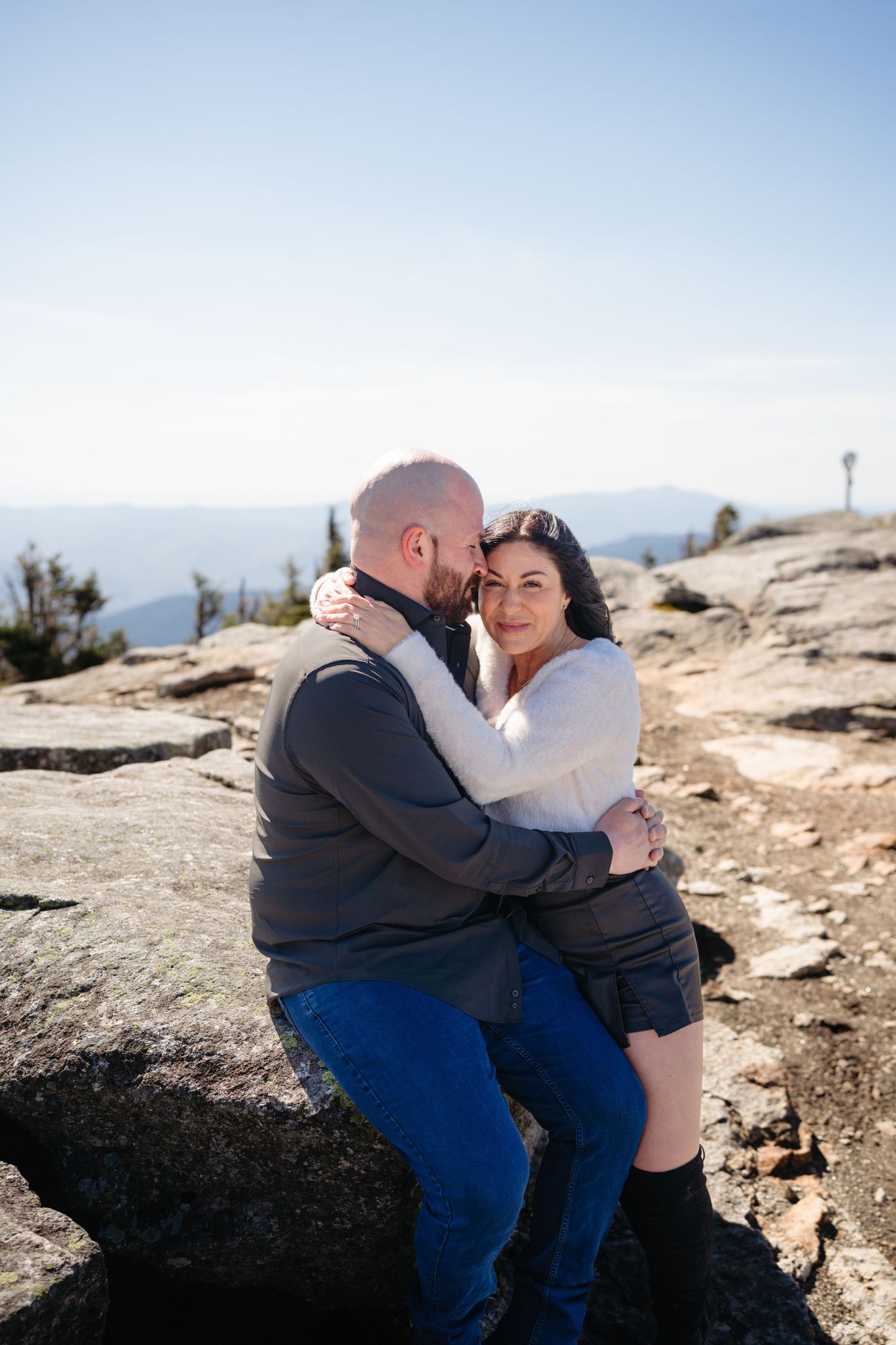 Couple embracing on rocky mountain summit, woman in white top smiling at camera while man kisses her cheek, scenic sky backdrop.